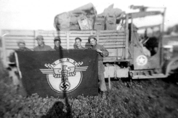 US soldiers standing beside an Army truck while holding aloft a captured Nazi flag.jpg